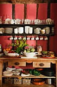 Gemüse und Obst in Schalen auf Holzanrichte unter Wandregal mit Geschirr an rot getönter Wand