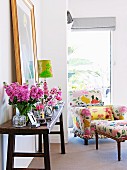 Rustic wall table with vases full of spring flowers; in the background a flower-patterned armchair with an ottoman