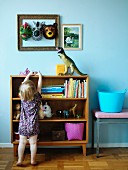 Little girl in front of shelves of toys; animal masks in picture frame and dinosaur figure against wall painted pale blue