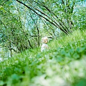 Kleines, blondes Mädchen, im Gras hockend; gekippter Horizont mit Bäumen