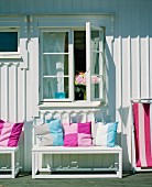 Colourful scatter cushions on bench outside white-painted wooden house on sunny day