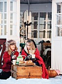 Zwei Mädchen mit Decke und Winterkleidung bei Teepause im Schnee vor einem Gartenpavillon
