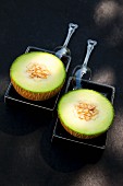 Two Galia melon halves in black ceramic dishes with plastic spoons