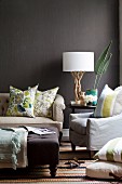 Upholstered seating with scatter cushions and table lamp with white fabric lampshade on natural wood base against wall painted dark brown