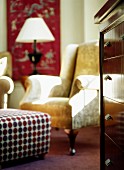 Patterned ottoman in front of antique leather armchair and chest of drawers in corner of living room