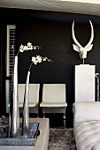 weiße Blume in Metallvase auf Tablett und weiße Sitzmöbel neben Tierkopf vor schwarzer Wand