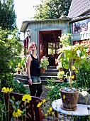 Lachende, junge Frau mit Pflanzentopf in sonnigem Garten vor nostalgischem Holzhaus