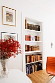 Branches of red flowers in vase next to fitted shelves of antiquarian books and partially visible leather chair