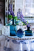 Blaue Glasvasen mit Lupinenblüten auf Konsolentisch mit Spitzenborte und gerafftem Stoff, darüber Antikspielge
