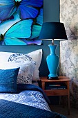 Bed with blankets, pillows and scatter cushions in shades of blue below picture of butterflies; blue bedside lamp on small cabinet