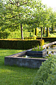 Geometrisch angelegte Hecken mit Bäumen, Rasenfläche und betoniertem Wasserlauf bilden gepflegte Gartenarchitektur