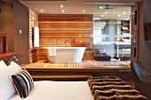 Vom Schlafzimmer aus zugängliches Designerbadezimmer mit freistehender weißer Badewanne vor holzverkleideter Wand