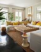 Großer gepolsterter Tisch inmitten weißer Polstersofas mit vielen gelben Kissen in hellem Wohnzimmer mit dunklem glänzendem Boden