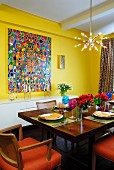 Esstisch aus Massivholz in gelbem Raum mit farbenfrohem, modernen Gemälde und darüberhängender, sternförmiger Hängelampe