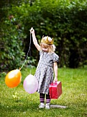 Little girl with crown, balloons & small suitcase in garden