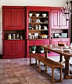 Country-house style kitchen-dining room with heavy wooden table and benches below wrought iron chandelier; matt red kitchen dresser in background