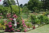 Bunt blühende Blumenrabatte in weitläufiger Gartenanlage; im Vordergrund rankende Rosen und eine Steinfigur