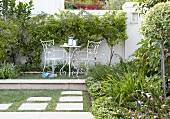 Gartenpodest mit filigranen Gartenmöbeln; dahinter eine weiße Gartenmauer mit blau blühenden Bäumchen