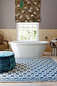 Grossräumiges Bad mit blauem Samt-Polsterhocker auf gemustertem Teppich vor Badewanne am Fenster