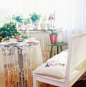 60s-style interior with bench & small table with lace tablecloth below window full of houseplants