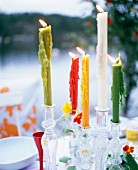 Candles of different colours in glass candlesticks on table in garden