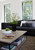 Square coffee table in front of black couch in living room with large windows; black vase of fresh wild flowers on table