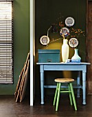 Pastel blue table and stool against olive green wall; collection of china plates on wall