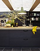 Black kitchen counter with stone top below glass ceiling; bouquet of olive branches on worksurface