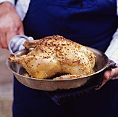 A person holding a pan with a crispy roast chicken