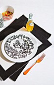 Black and white patterned plate and bottle of orangeade on black and white place mat with orange fork to one side