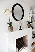 White potted orchids and ornament under glass cover on mantelpiece below round mirror with black frame on wall