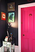 Shiny table lamp illuminating collection of pictures on dark grey wall next to hot pink panelled door