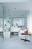 Man sitting on classic La Chaise chair in front of modern, glass bathroom area with marble washstand and mirrored cabinet