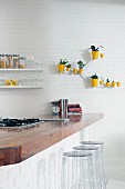 Solid wood kitchen counter and plexiglass bar stools in front of white brick wall; arrangement of sunshine yellow planters on wall