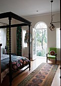 Black four-poster bed with artistically turned bedposts in bedroom with open balcony door and view of garden