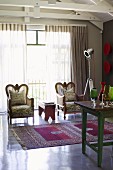 Königliche Sessel vor großer Glasfront in Wohnzimmer mit edlem Teppich, Vintage Tisch mit darauf stehender Gläsersammlung und einer Filmlampe