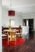 Weitläufiger Raum mit Essgruppe aus hellem Holz auf rotem, runden Teppich; im Hintergrund ein breiter Durchgang ins Wohnzimmer