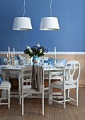 Festlich gedeckter weißer Esstisch mit Silberkerzenleuchtern vor blauer Wand und zwei modernen Pendelleuchten