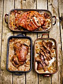 Three different roasted joints of meat in roasting tins on a wooden surface