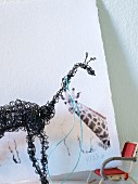Black, wire giraffe sculpture in front of picture of giraffe on white background