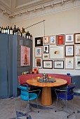 Runder Holztisch im Biedermeierstil und filigrane Metallstühle vor Wand mit Bildersammlung, in Zimmerecke blauer Metallschrank mit oben abgestellten Flaschen