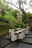 Stühle mit weisser Husse um Holztisch auf Terrassen Platz in tropischem Garten