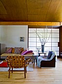 Holzstuhl und Sessel neben gemütlichem Sofa mit Kissen vor Schiebeelement mit japanischem Papier bespannt in modernem Wohnraum mit holzverkleideter Tonnendecke