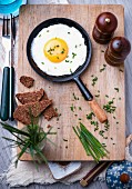 A fried egg with chives in a frying pan, wholegrain bread, salt and black pepper