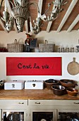 White enamel bread bins and ceramic pots on rustic kitchen worksurface; aphorism written on red paper