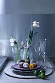 Still-life arrangement with decorative Easter eggs on dishes, glasses and spring flowers on grey concrete surface against grey-painted wall