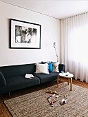 Toys on rug in front of black sofa below photo on wall and round side table in front of transparent curtains on window