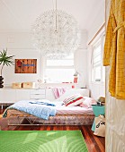 Green rug on wooden floor, bed with retro metal frame and scatter cushions, spherical lamp with many flower-shaped elements and white sideboard in background