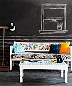 White-painted wooden bench and table with colourful mixture of textiles; wall and floor painted with blackboard paint