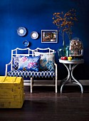 White wicker bench with floral seat cushion and scatter cushions next to glass vase of flowers on round side table against blue-painted wall; yellow wooden trunk in foreground
